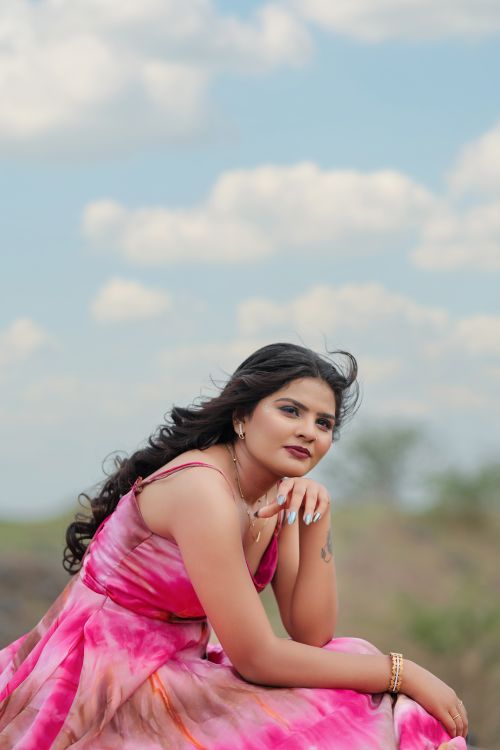 Free Elegant woman in a flowing pink dress posing outdoors, with clouds and nature in the background. Stock Photo
