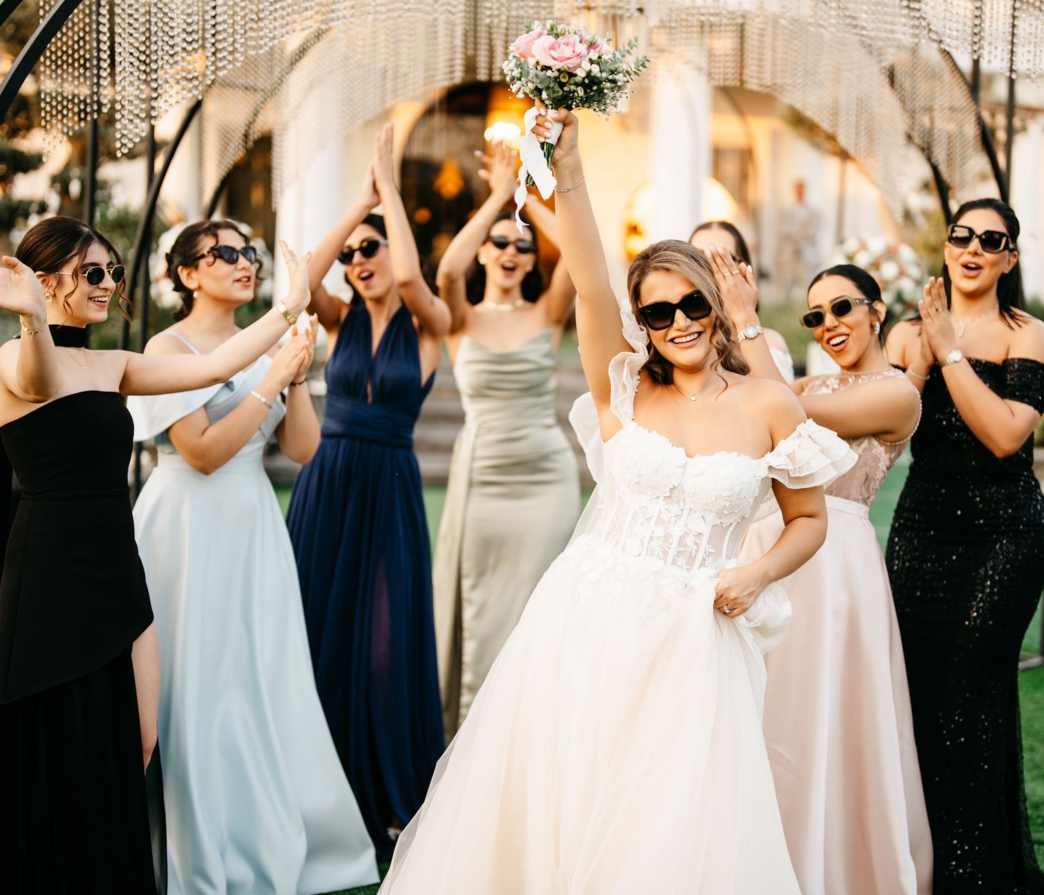 Bride and bridesmaids celebrating outdoors in a joyful wedding scene with sunglasses and dresses.
