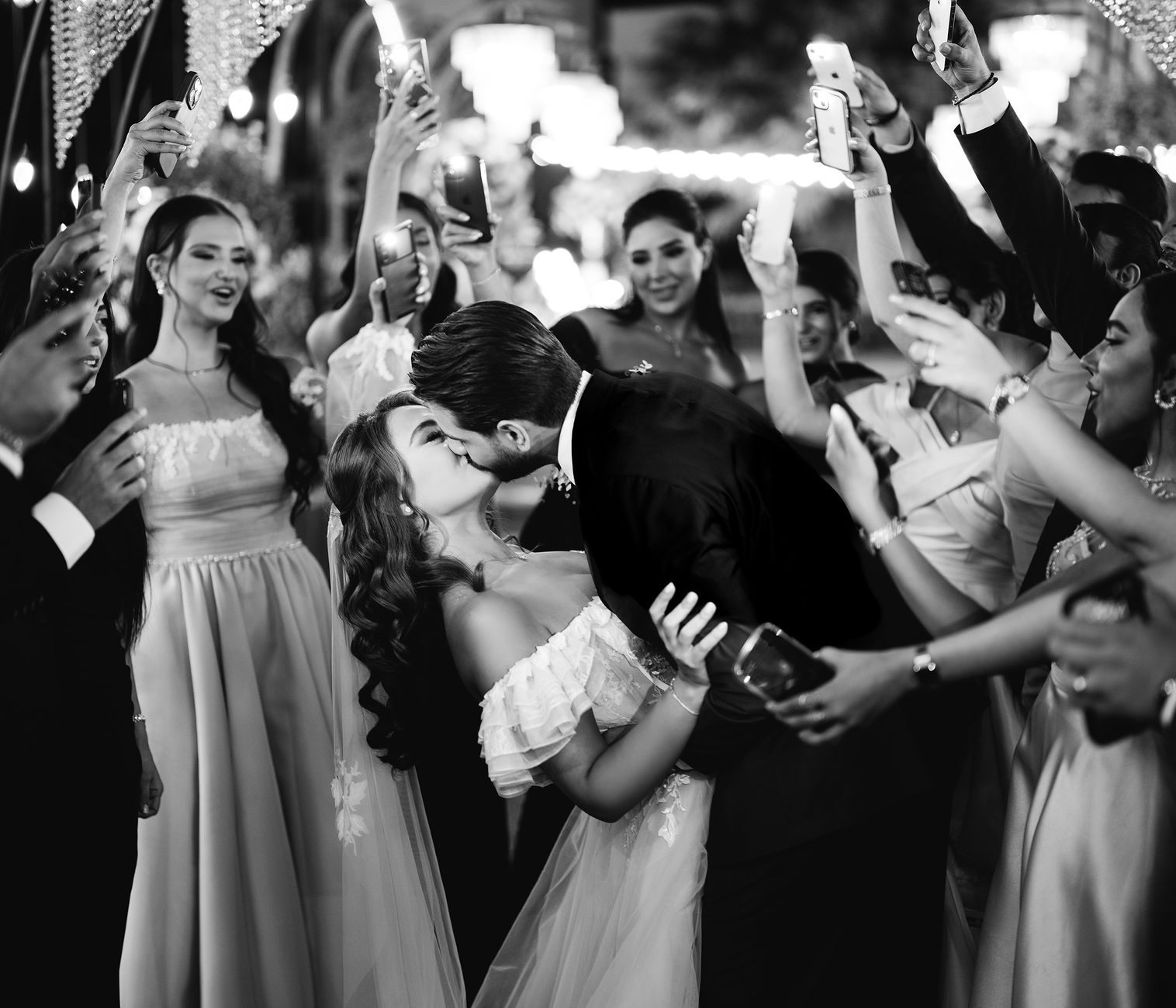 A newlywed couple shares a romantic kiss, surrounded by celebrating guests capturing the moment.