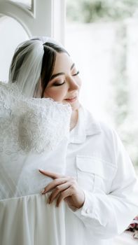A joyous bride in white holding her wedding dress, exuding happiness and anticipation indoors. - Fotografer Sidoarjo