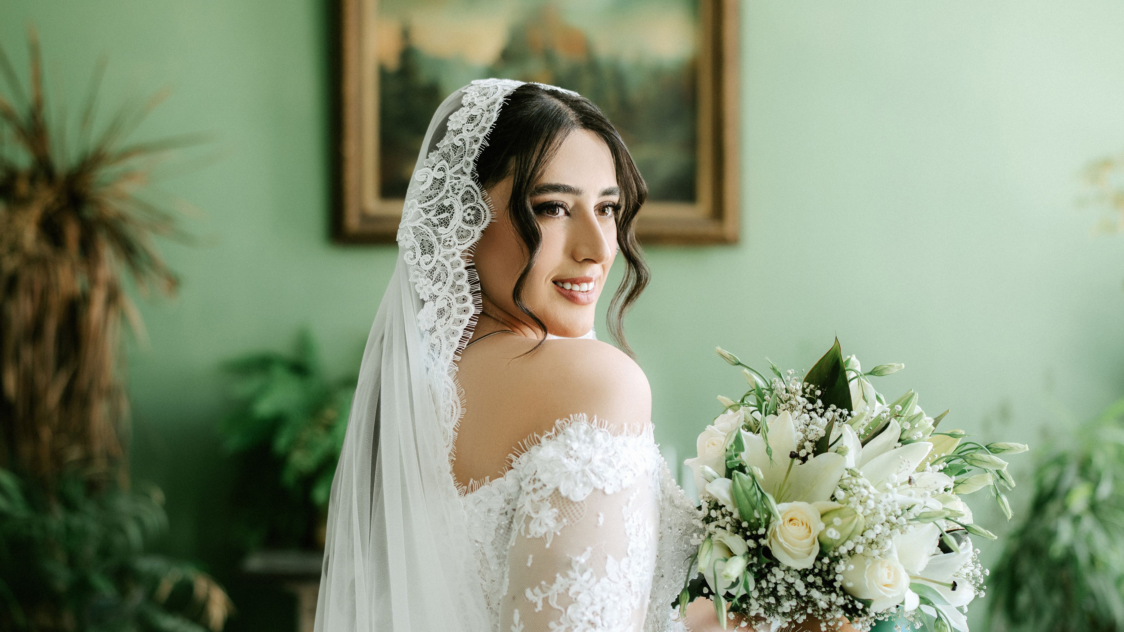Happy bride posing with floral bouquet indoors, showcasing her elegant lace wedding dress.
