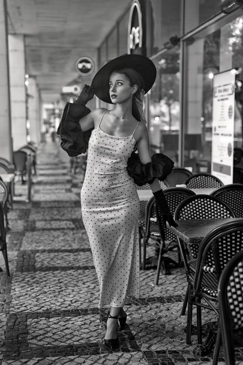 Free Stylish woman in polka dot dress and hat walks through an outdoor cafe seating area in black and white. Stock Photo