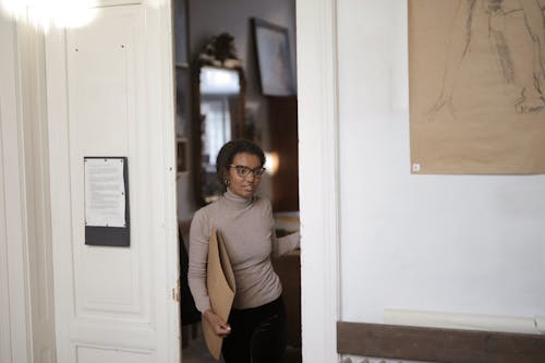 Free Young African American female student in casual clothes and eyeglasses leaving classroom and carrying folder with pictures during studying in art college Stock Photo