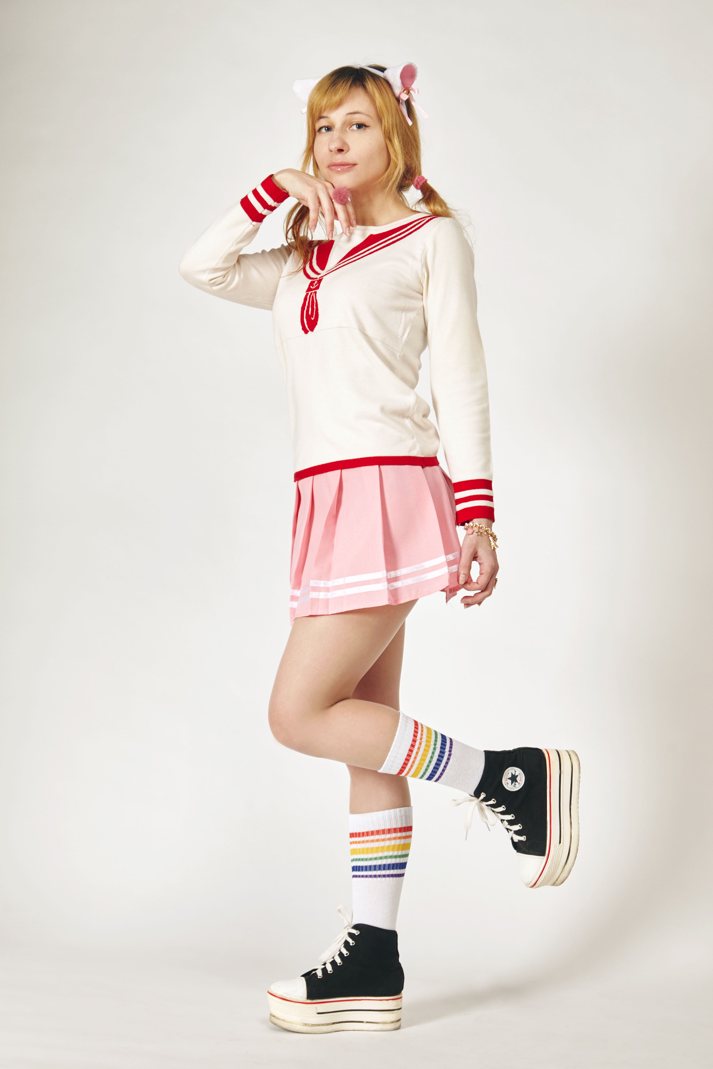 Side view of young feminine slender woman in stylish platform footwear and striped knee socks touching chin while standing on one leg and looking at camera near white wall