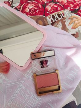 Elegant blush and compact mirror on a pink fabric, with floral and magazine backdrop.