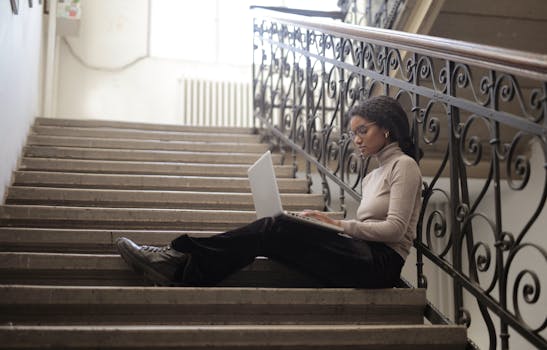 Young woman using laptop on indoor staircase, embracing remote work lifestyle.