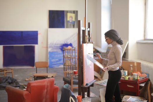 Side view of young black female artist in casual clothes standing near easel and drawing picture while working in studio