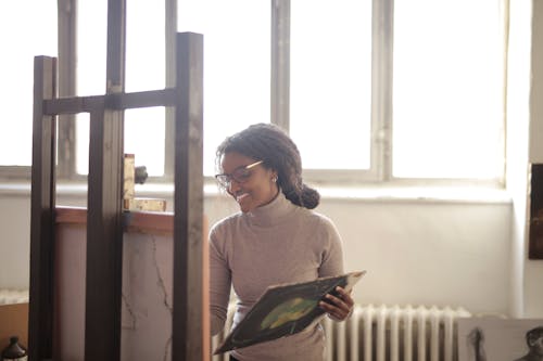 Positive smiling young black woman in casual wear standing on background of window and drawing picture on easel in light workspace