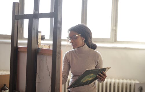 Positive young African American lady in beige turtleneck and eyeglasses standing near easel against bright window and drawing picture in creative studio