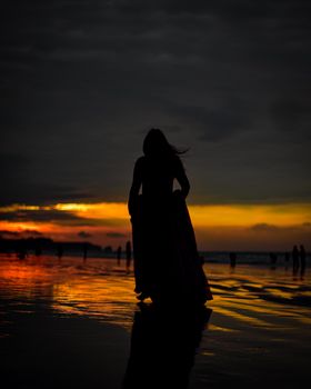 A silhouetted woman stands on a beach during a vibrant sunset, reflecting on wet sand.