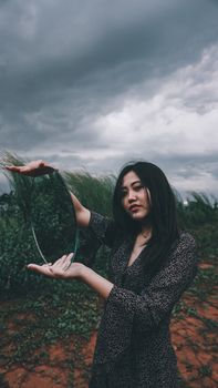 一位身着民族服饰的年轻女士站在干燥的小路上，傍晚时分，她望向镜头，身旁杂草丛生的草地上有一面椭圆形的镜子，映照着多云的天空