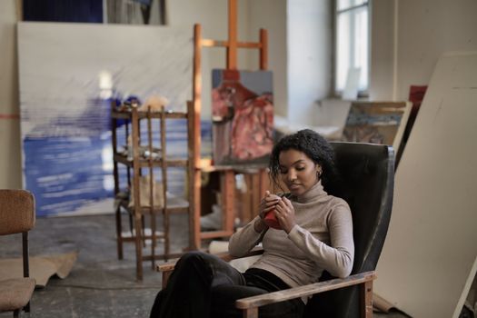 Relaxed black lady in casual wear sitting on shabby black armchair drinking hot beverage and looking down while resting in modern art studio