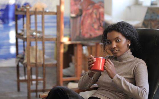 A woman enjoying a drink in an art studio, holding a red mug and sitting comfortably.