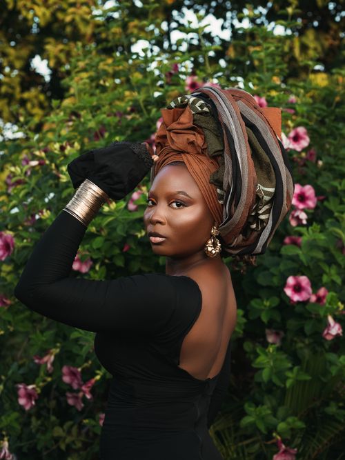 Free A beautiful woman adorned with an African headwrap poses gracefully amid blooming flowers in a lush garden setting. Stock Photo