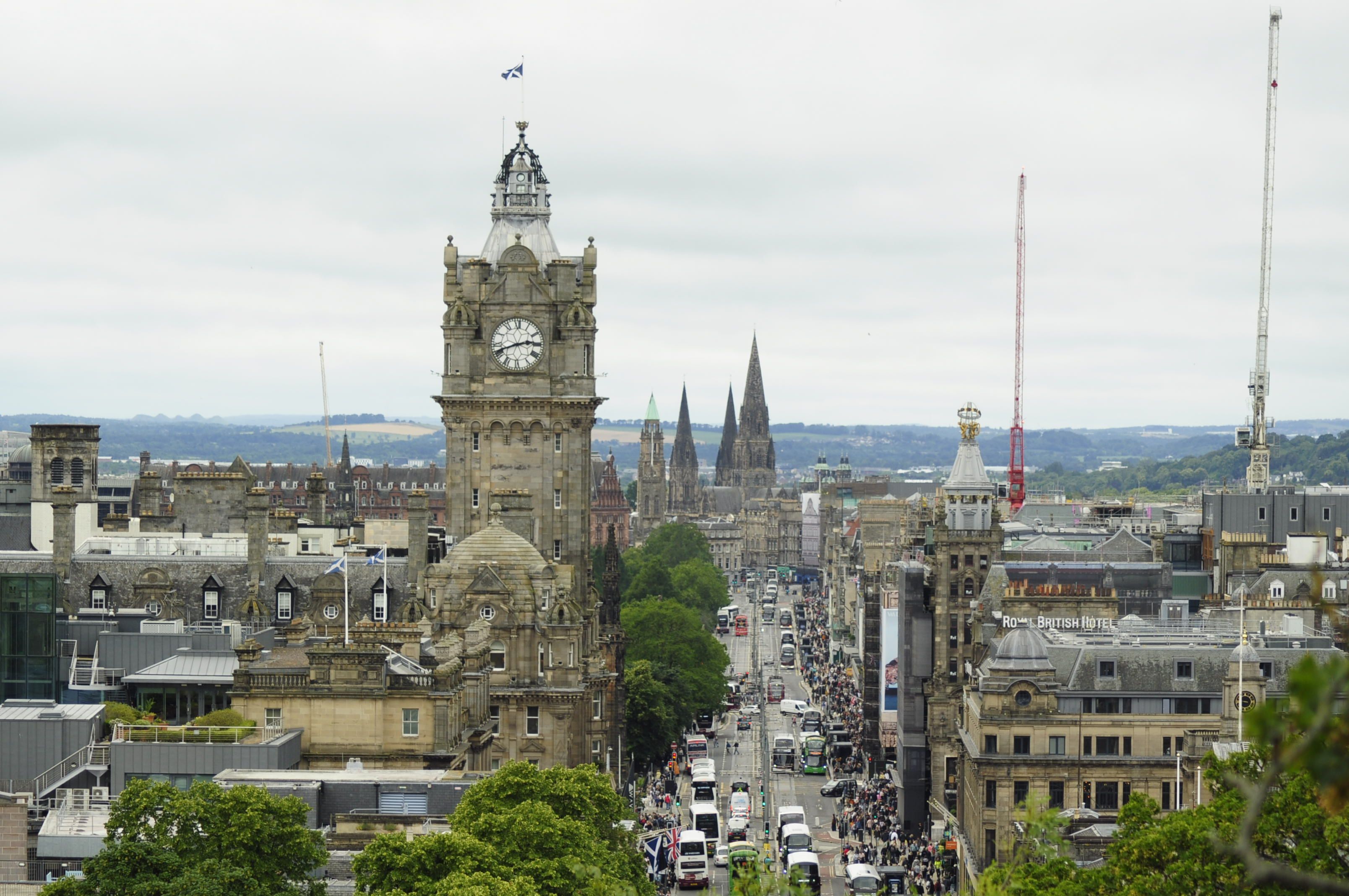 A stone clock tower rising above a busy street seen from high up, with Gothic spires in the distance under an overcast sky