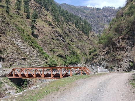 A scenic view of a rustic red bridge in a lush mountain valley with a dirt road.