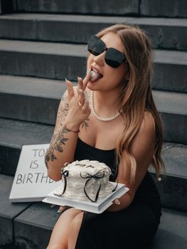 A stylish young woman enjoys her birthday with elegance, holding a white cake while sitting on stone steps.