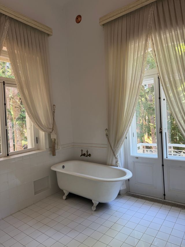 A clawfoot tub on white tiles under tall sunlit windows with linen curtains