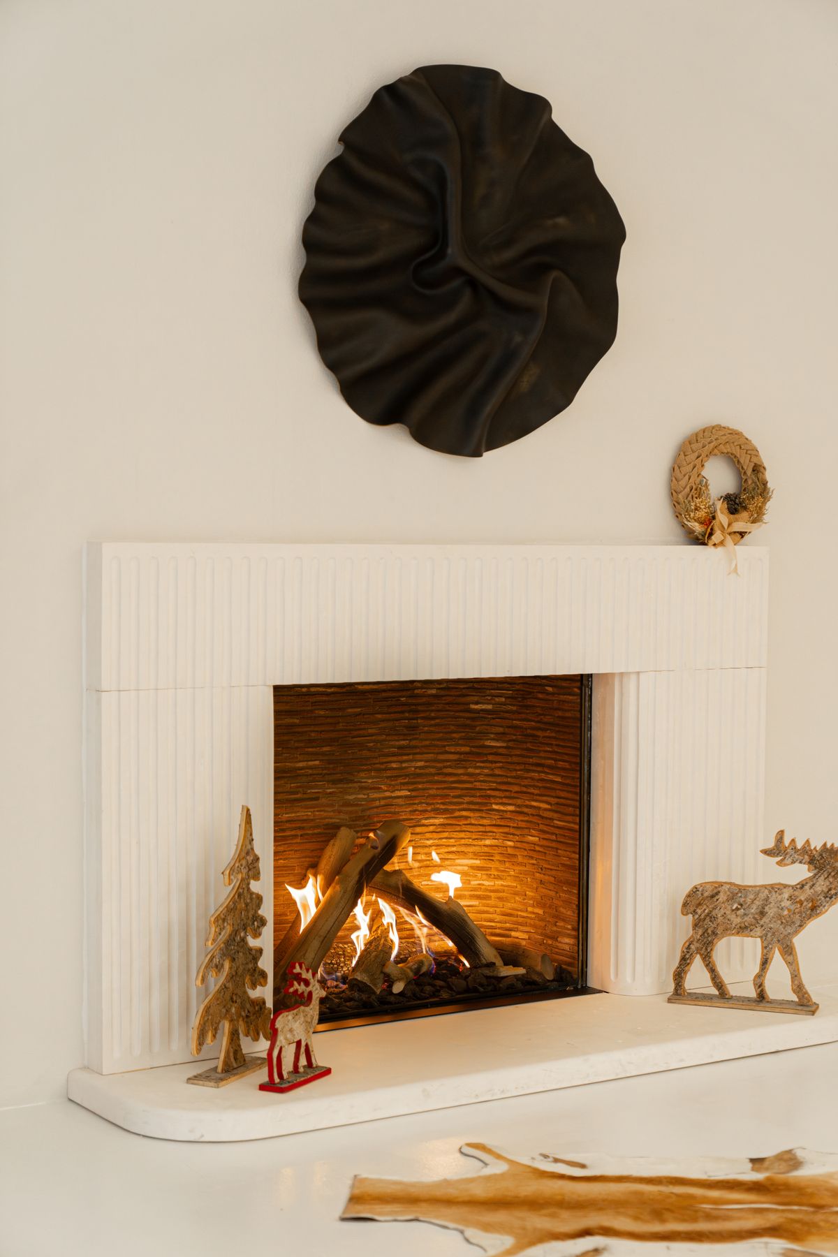 Minimal white fireplace with a lit fire, a small wooden Christmas tree and a wooden deer figurine on the hearth