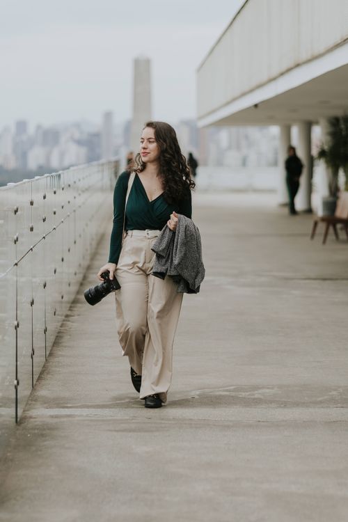 Free Confident female photographer walking with a camera on an urban rooftop terrace. City skyline in the background. Stock Photo