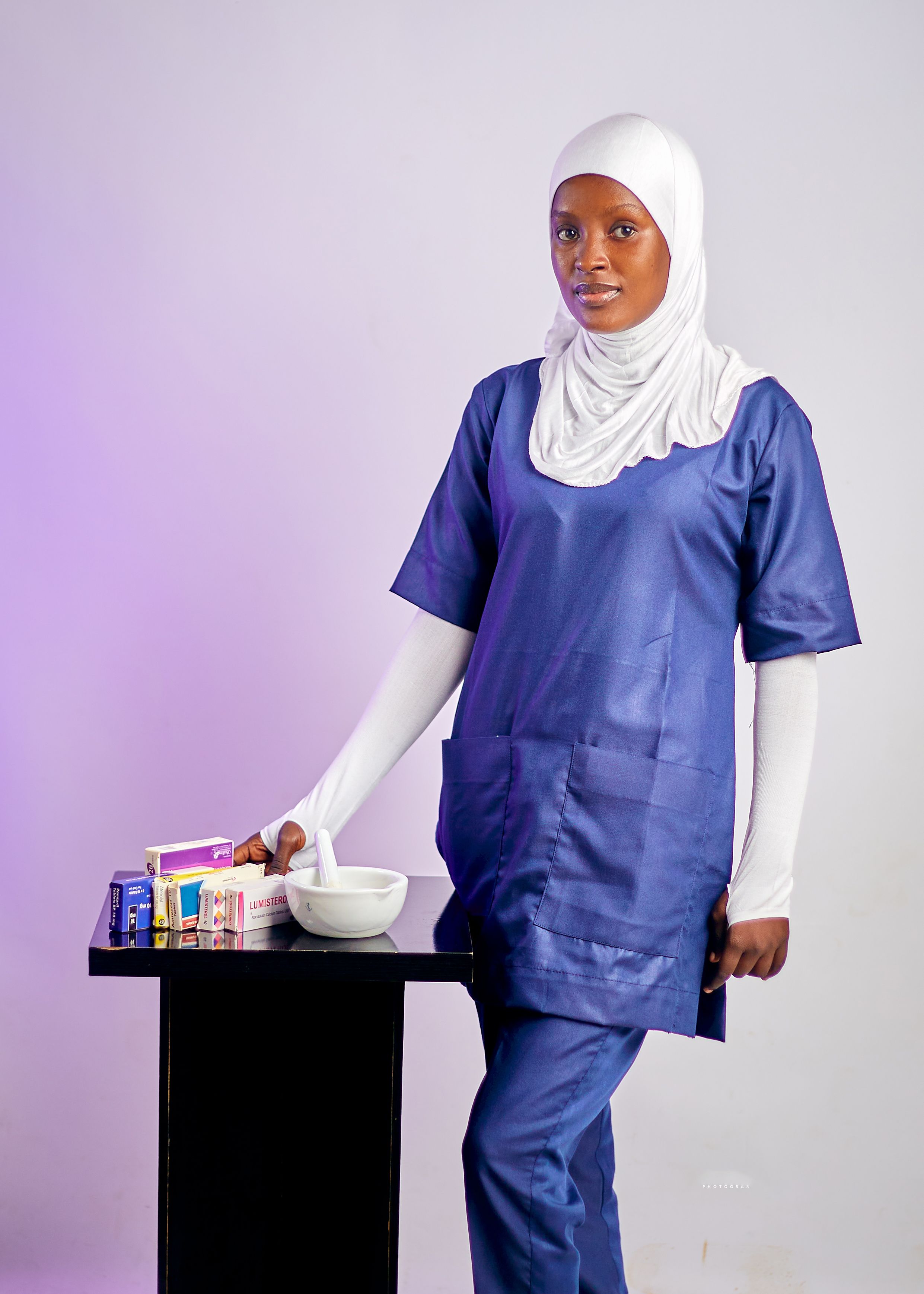 African female healthcare professional in scrubs and hijab beside medicine table indoors.