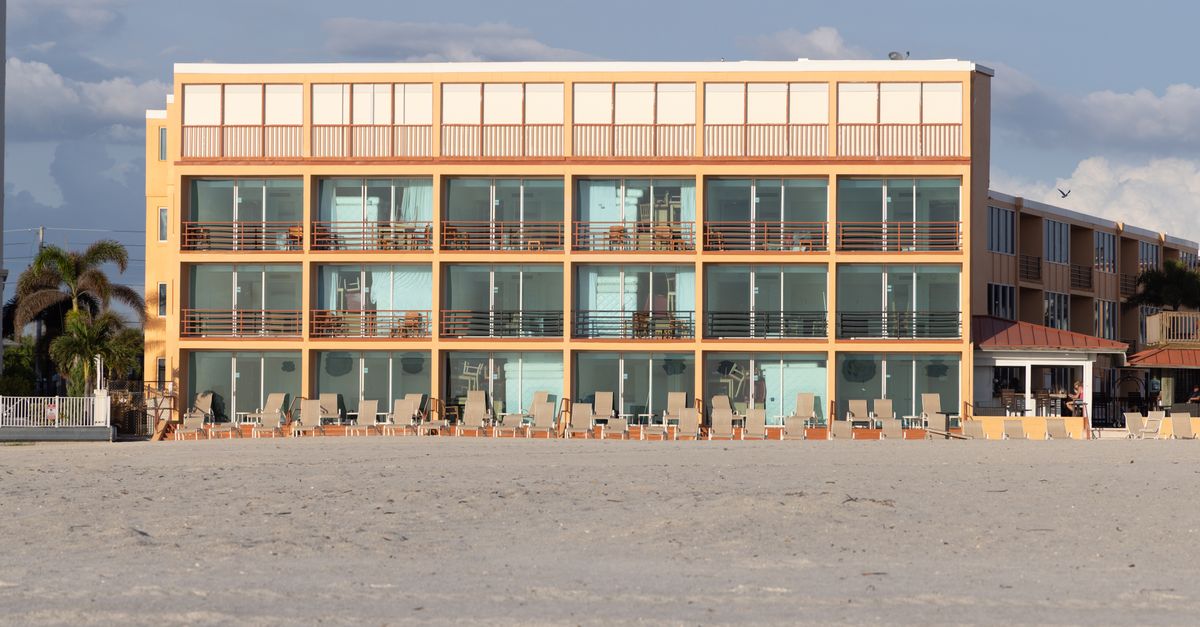 A modern beachfront hotel with balconies, situated on a sandy beach under a clear blue sky.