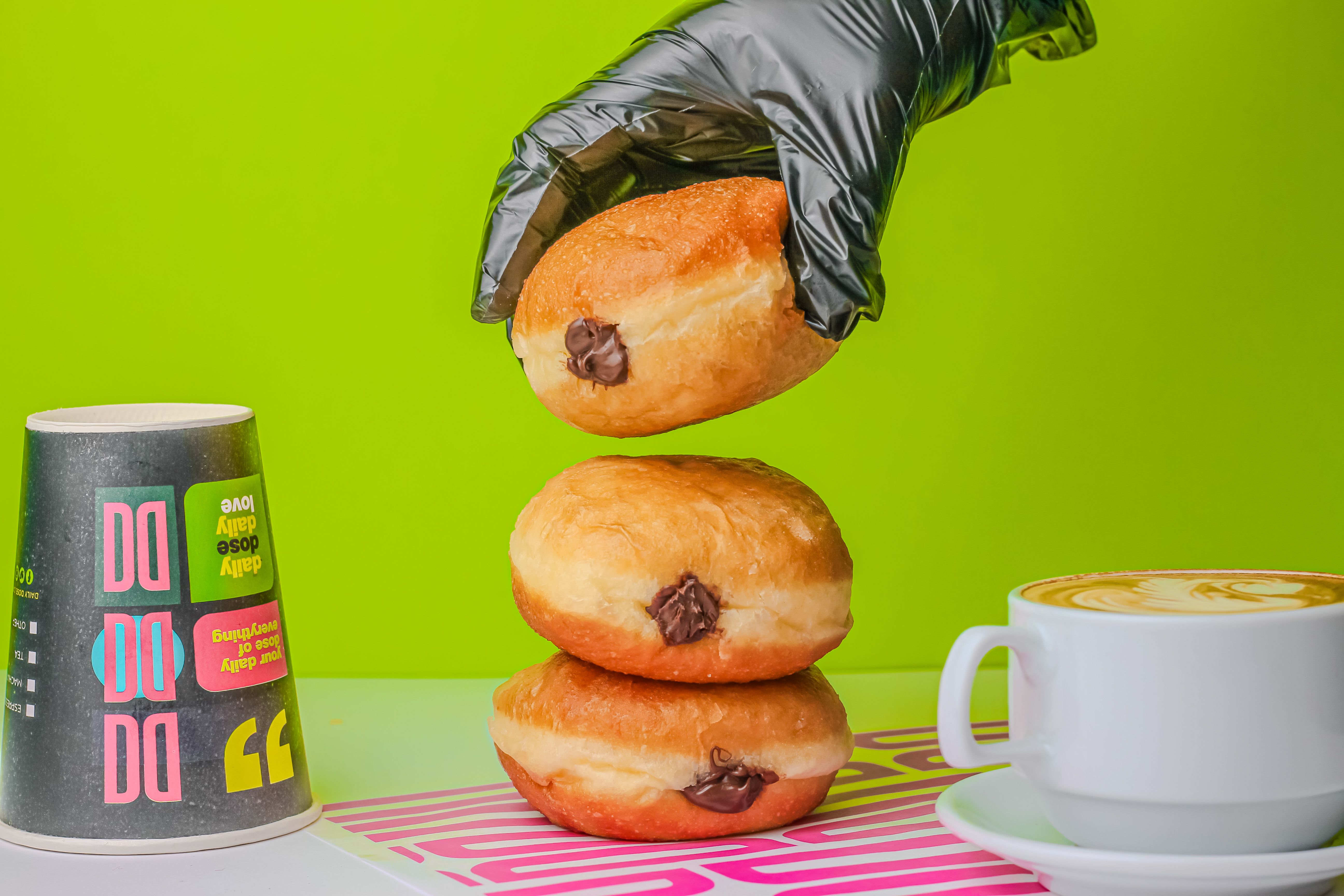 A stacked donut with chocolate filling beside a cappuccino and colorful cup on a vibrant green backdrop.