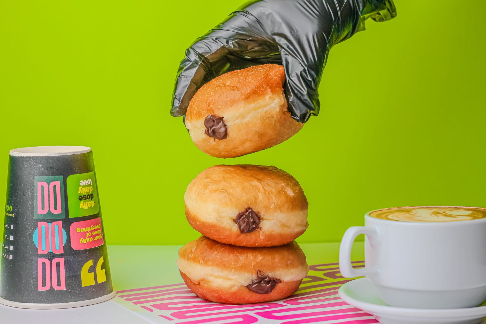A stacked donut with chocolate filling beside a cappuccino and colorful cup on a vibrant green backdrop.