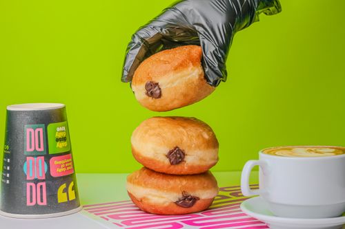 Free A stacked donut with chocolate filling beside a cappuccino and colorful cup on a vibrant green backdrop. Stock Photo