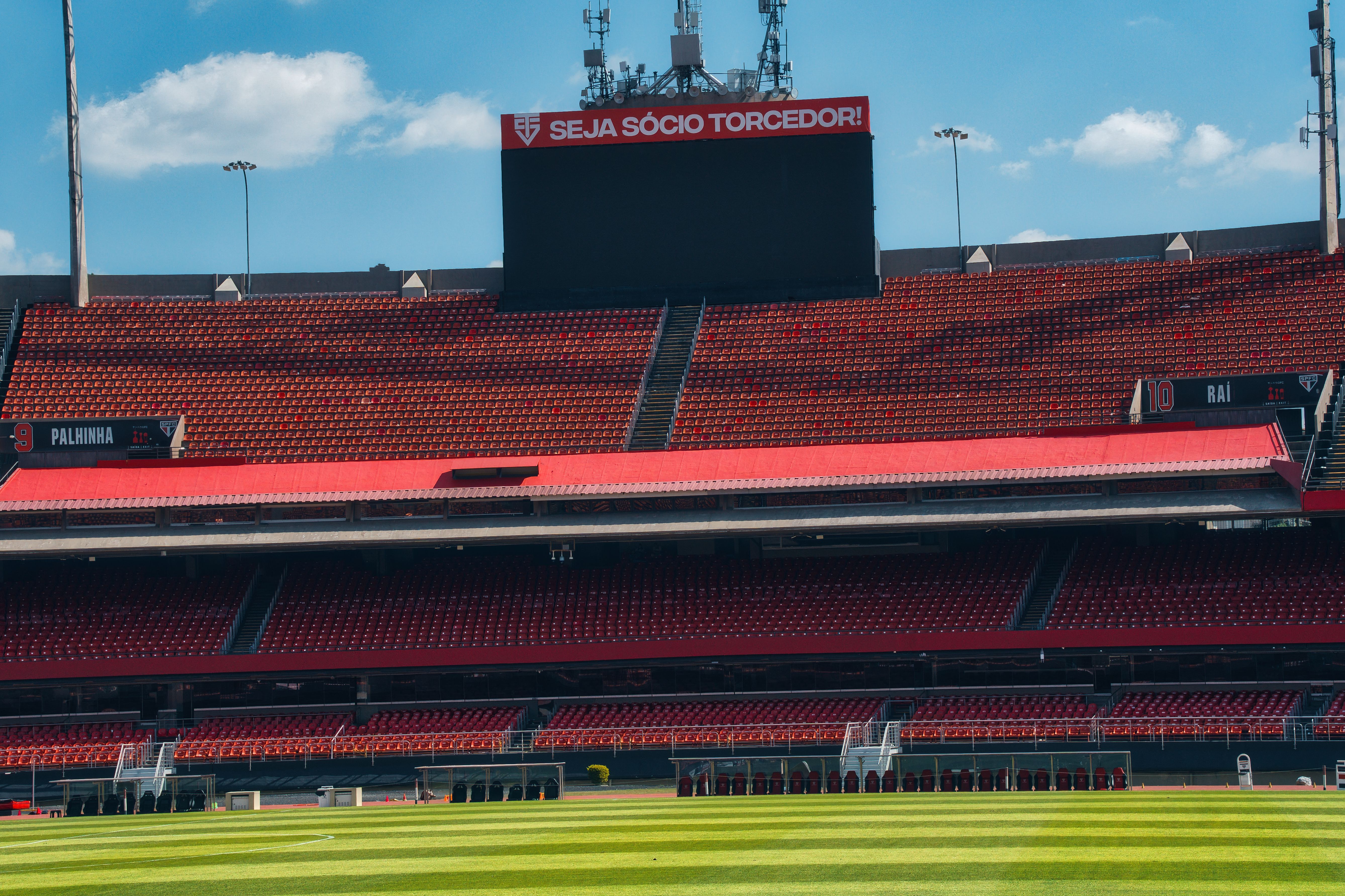 Empty football stadium with red stands, scoreboard, and lush green field under a bright sky.