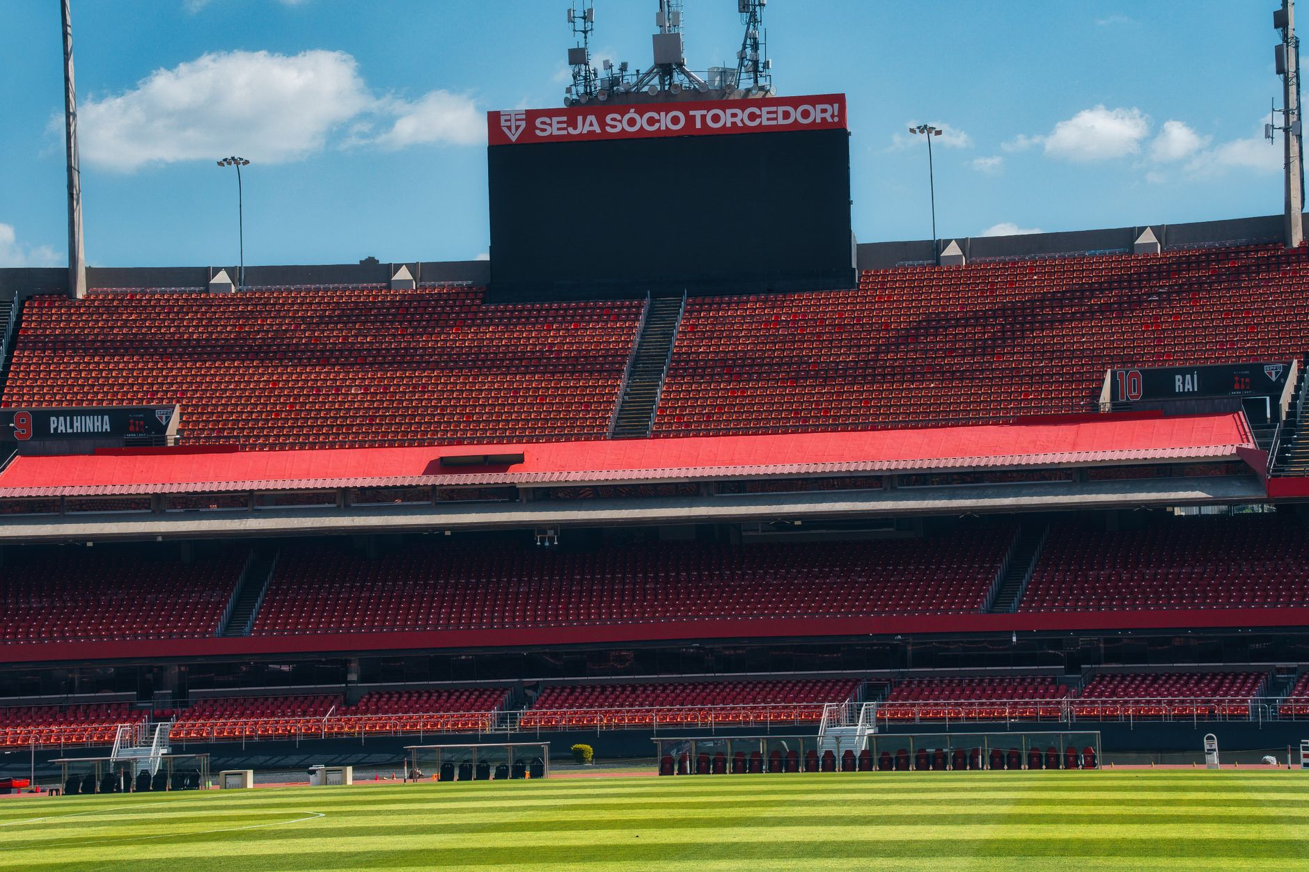 Empty football stadium with red stands, scoreboard, and lush green field under a bright sky.