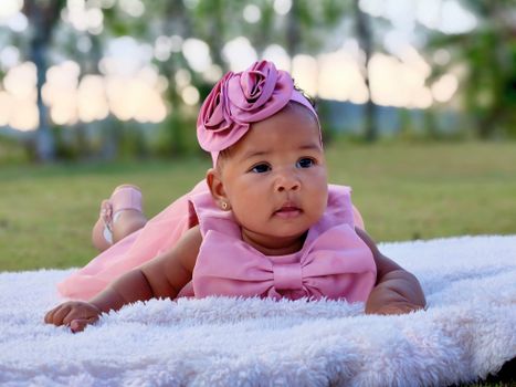 Cute baby girl lying on a blanket outdoors in a pink dress during the day.