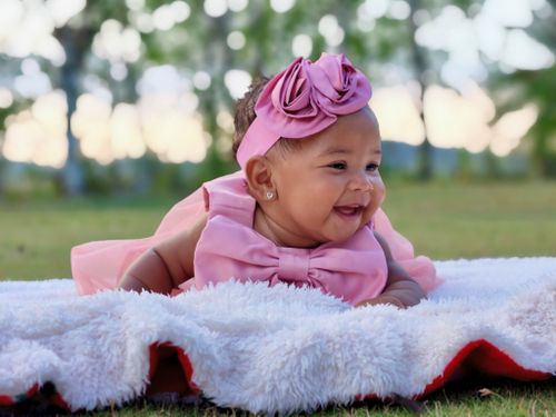 Free Charming photo of a baby in a pink dress smiling, lying on a soft blanket outdoors. Stock Photo