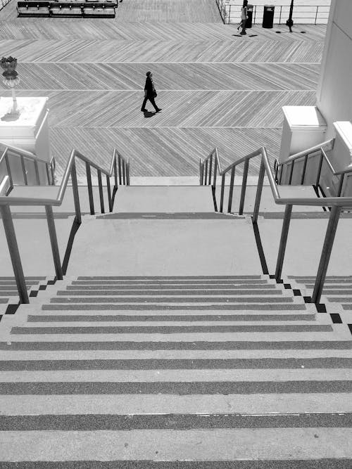 From above side view black and white of silhouette of anonymous male strolling on pavement near concrete staircase with horizontal lines on steps and metal fence with handrails