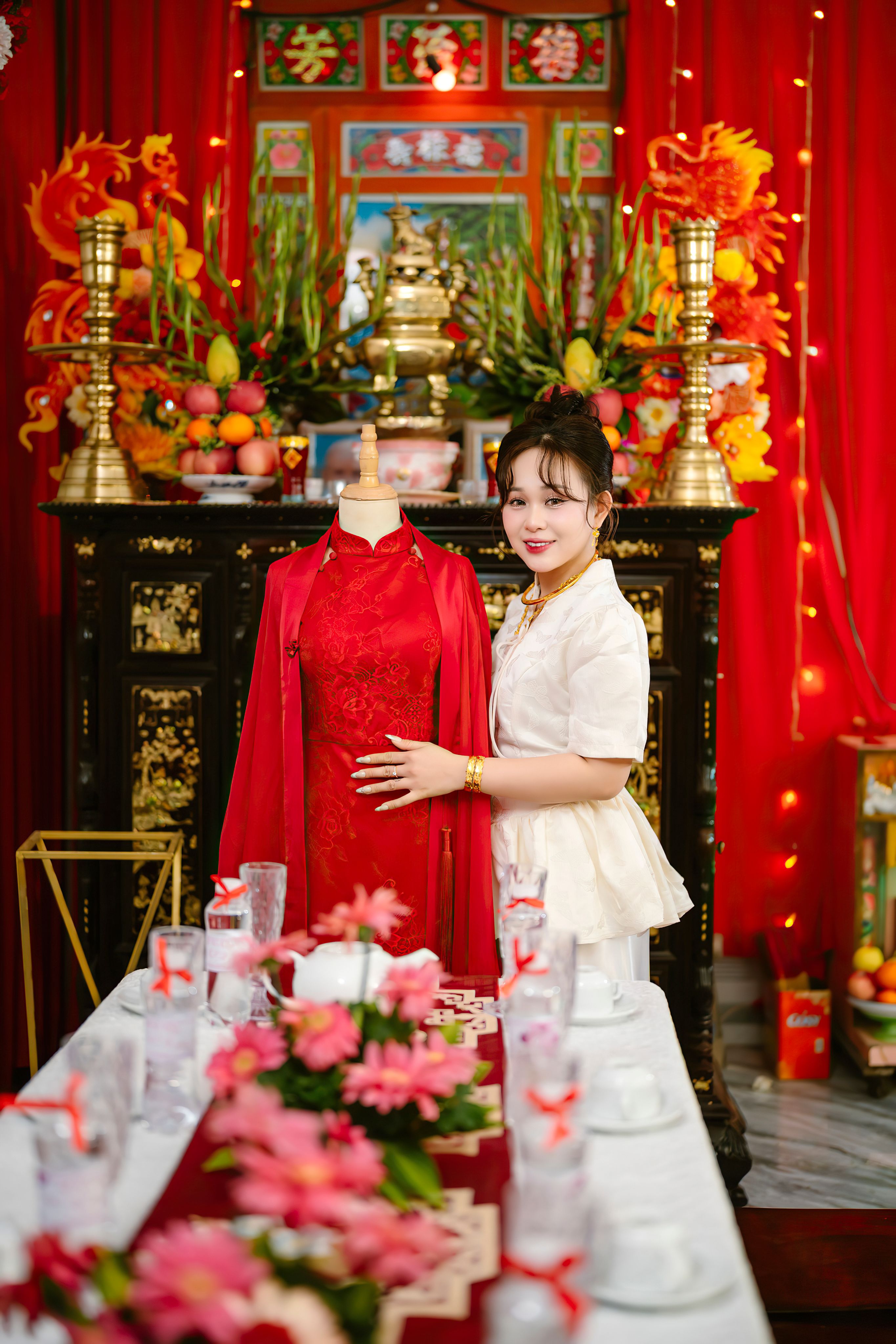 A woman stands beside a traditional Vietnamese wedding dress in a brightly decorated room.