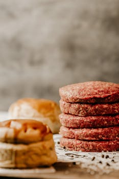 Stack of raw beef patties on a rustic table with buns, ideal for burger preparation theme.