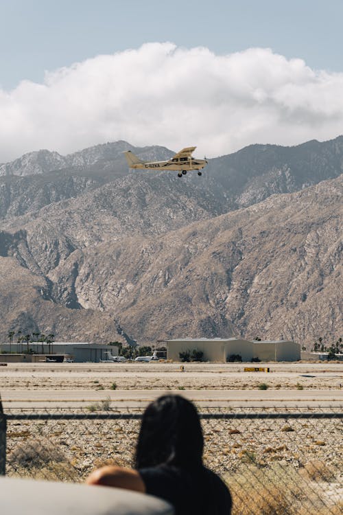 Back view of crop anonymous female standing near fence and roadway under flying helicopter near mounts and sky with thick clouds in daylight