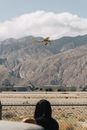 Back view of crop anonymous female standing near fence and roadway under flying helicopter near mounts and sky with thick clouds in daylight
