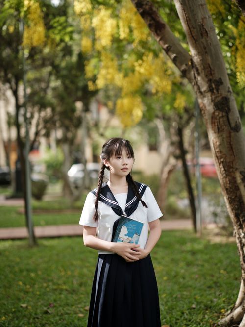 Free A young woman in a school uniform stands thoughtfully holding a book outdoors in a sunny garden. Stock Photo
