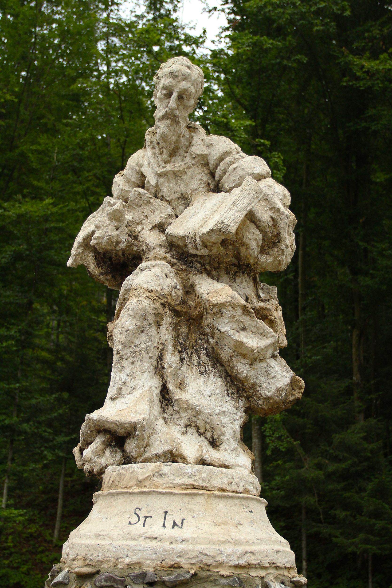 A weathered stone statue of a saint in a lush forest backdrop.