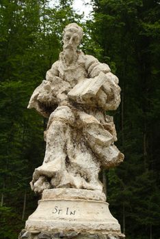 A weathered stone statue of a saint in a lush forest backdrop.