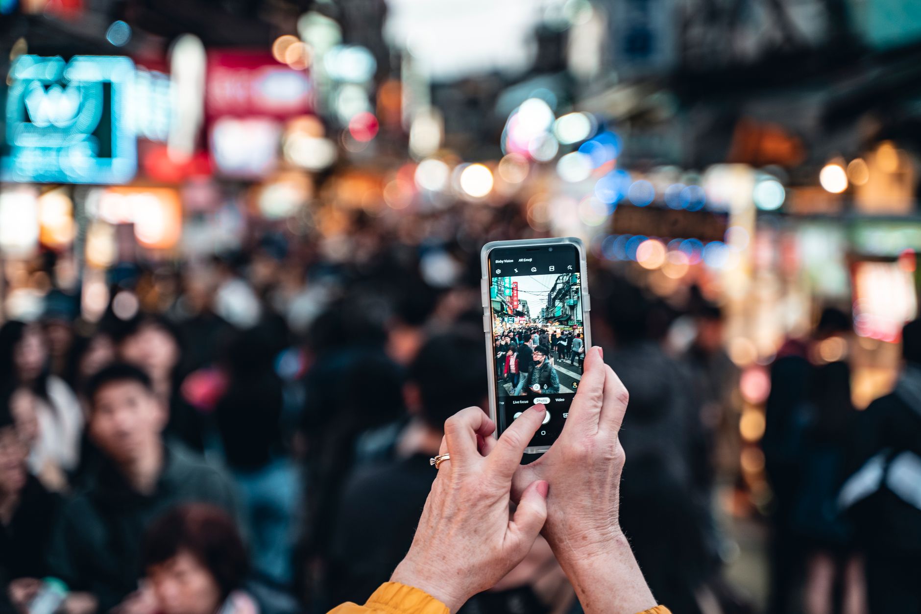 Hands holding a smartphone, capturing a bustling city street filled with people, vibrant signs, and lively activity.