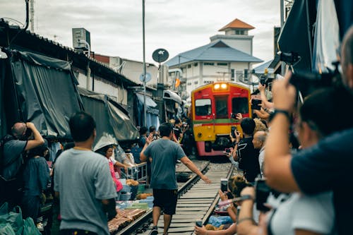 Back view of anonymous ethnic men and women standing near railway while taking photo on cellphones of old colorful tram on narrow street behind building under cloudy sky