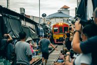 Back view of anonymous ethnic men and women standing near railway while taking photo on cellphones of old colorful tram on narrow street behind building under cloudy sky