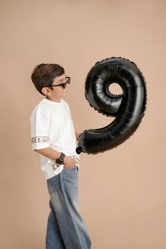 Young boy with glasses holding a black inflatable number 9 balloon against a beige backdrop.