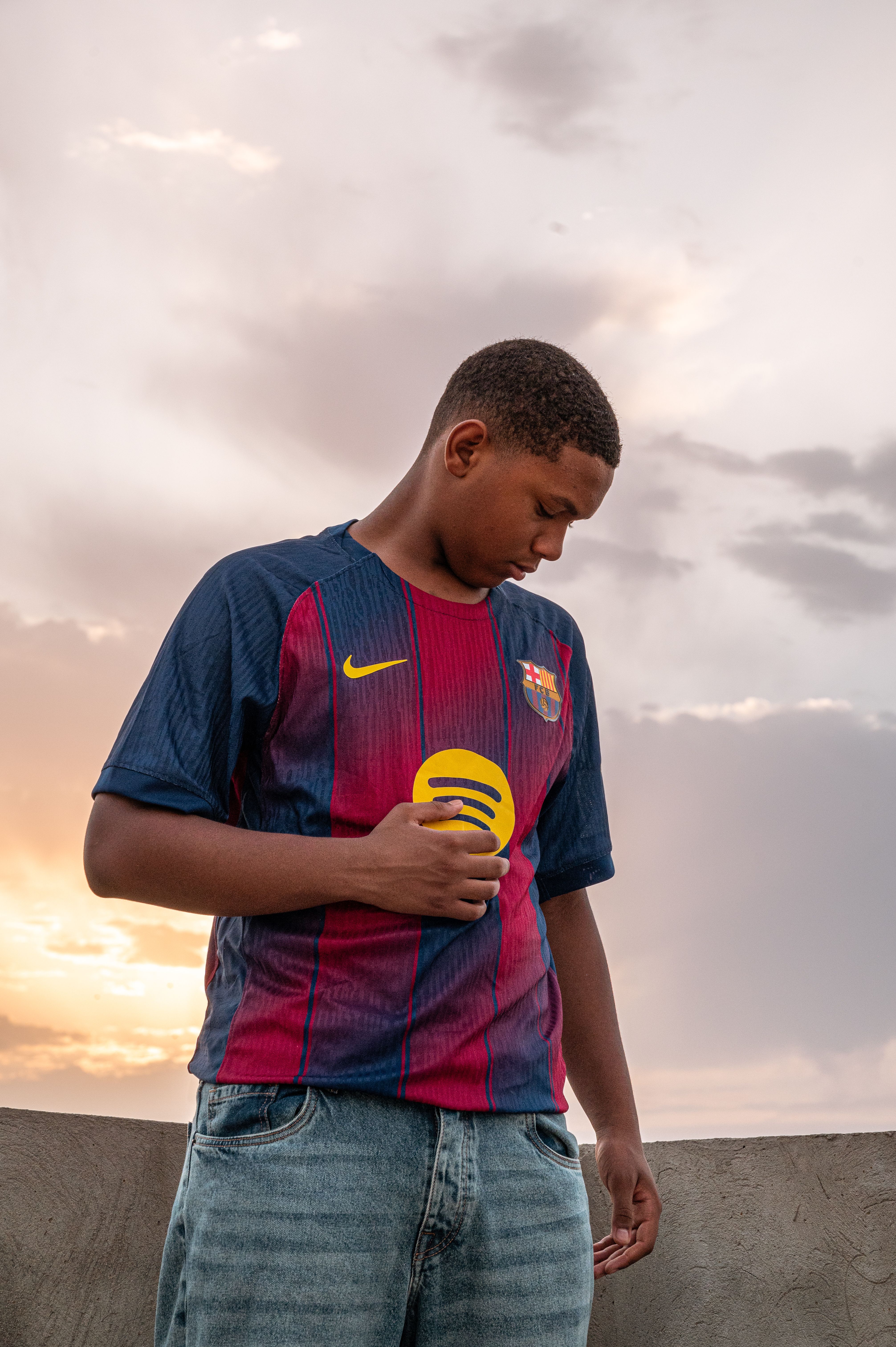 Young man in sports jersey looks down against a sunset backdrop.