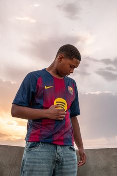 Young man in sports jersey looks down against a sunset backdrop.
