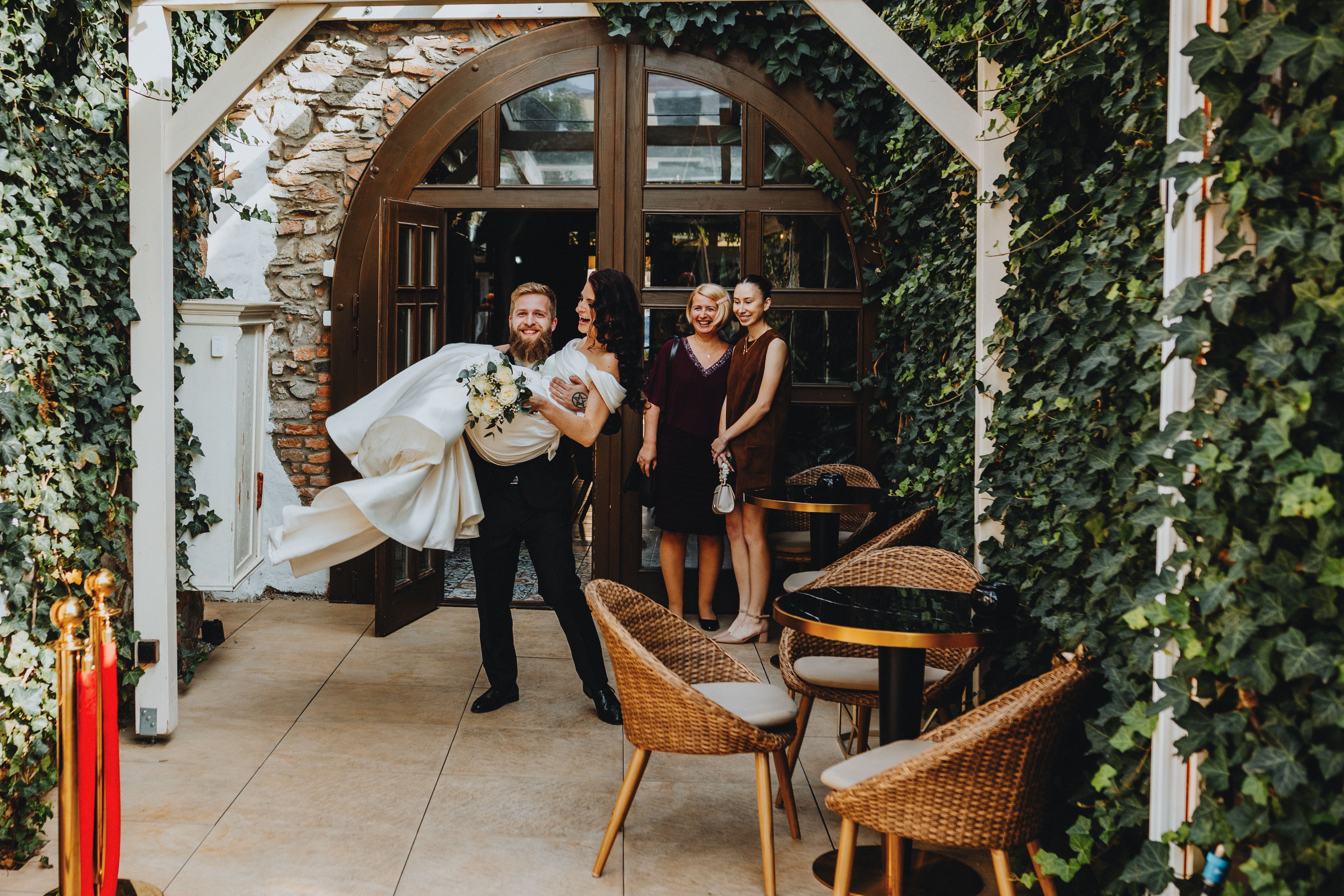 Bride being joyfully carried by the groom, surrounded by friends at an elegant outdoor wedding reception.
