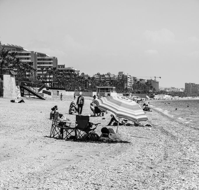 Imagen en monocromo de una playa de guijarros con sombrillas y edificios distantes.