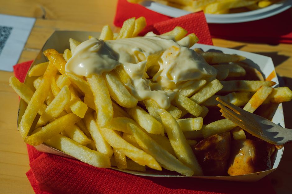 Plate of currywurst with fries and mayo, served outdoors with vibrant lighting.